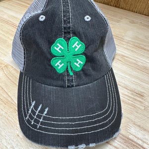 Puede incluir: Una gorra de béisbol negra y verde bordada con el logotipo de 4-H, con un aspecto desgastado. La gorra tiene una parte trasera de malla y una visera blanca.