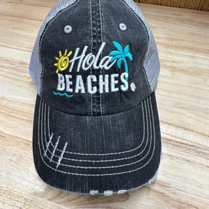 Op de afbeelding: Een zwarte en grijze distressed trucker cap met een wit geborduurd ontwerp dat "Hola Beaches" zegt met een zon, palmboom en golfgrafiek.