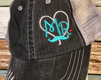 May include: A black and gray trucker hat with a distressed denim front and a mesh back. The hat has a turquoise embroidered design of a heart with the word "MR" inside and a mustache underneath.