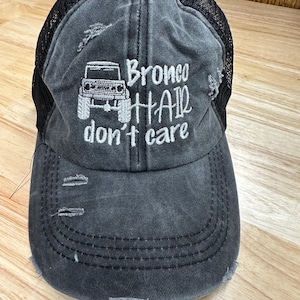 Könnte beinhalten: Verwaschene graue Baseballkappe mit schwarzem Mesh-Rücken. Die Kappe hat den gestickten Text "Bronco Hair don't care" mit einer Grafik eines Bronco-Fahrzeugs in Weiß. Die Kappe hat einen vorgekrümmten Schirm und einen Used-Look.