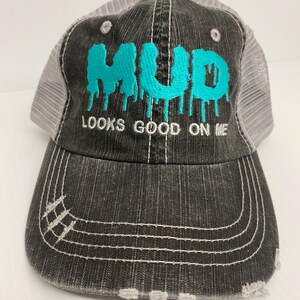 Puede incluir: Una gorra de camionero negra y gris con una malla blanca en la parte posterior. La parte delantera de la gorra tiene un diseño bordado en azul turquesa que dice "MUD LOOKS GOOD ON ME".