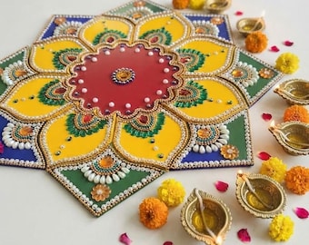 May include: A decorative mandala-style rangoli floor art piece featuring a central red circle surrounded by yellow petals and green triangular corners, embellished with white pearls, colorful beads, and rhinestones, accompanied by small lit oil lamps.