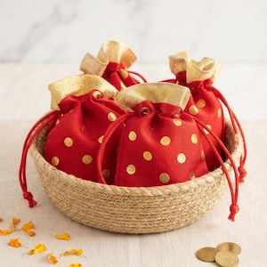 May include: Four small red fabric drawstring pouches with gold polka dots and gold trim, gathered at the top with red cords. The pouches are arranged inside a round, woven tan basket. Scattered nearby are small yellow flower petals and gold coins.
