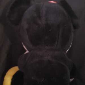 May include: Black plush toy with a yellow tail and a pink bow. The toy is shaped like a dog with large ears.