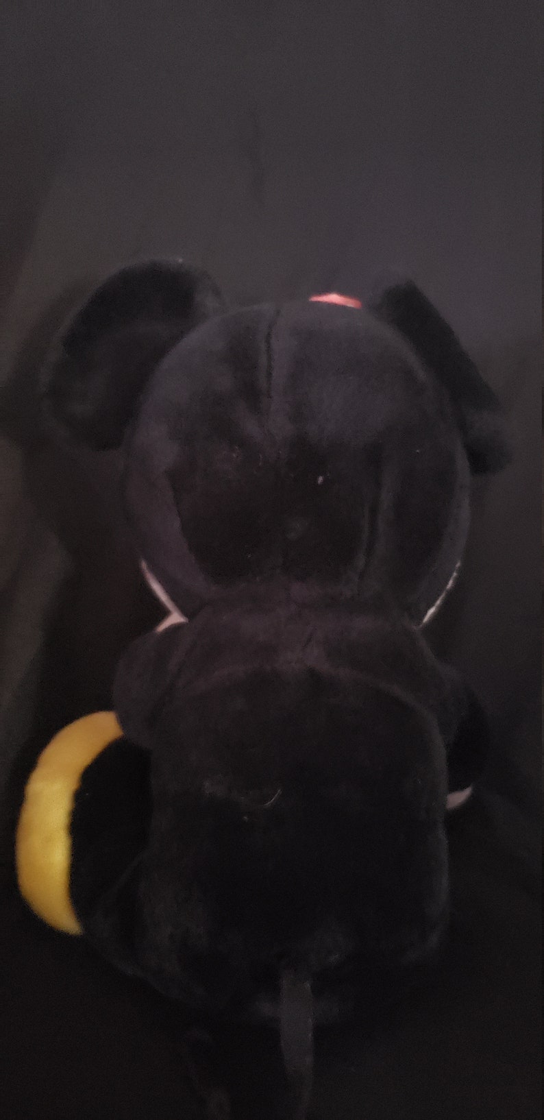 May include: Black plush toy with a yellow tail and a pink bow. The toy is shaped like a dog with large ears.