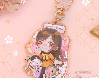 May include: A gold-coloured acrylic charm featuring a cartoon girl with brown hair and a bow, giving a thumbs-up. The charm includes two small doll figures and the text "A dream of Spirutt's Artwork and Store". The charm is attached to a gold-coloured clasp.