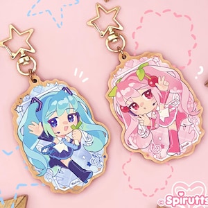 Op de afbeelding: Twee houten sleutelhangers in anime-stijl met gouden stervormige sluitingen. Eén heeft een blauwharig personage, de andere een rozeharig personage. Beide hebben gedetailleerde illustraties en muzieknootaccenten. Op een roze achtergrond.