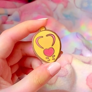 May include: A yellow enamel pin shaped like a lightbulb with a pink heart inside. The heart has a sparkly texture.