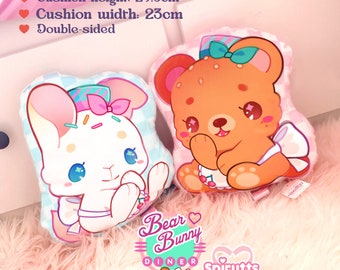 May include: Two double-sided plush cushions. One features a white bunny with blue eyes and a pink bow, the other a brown bear with a bow. The cushions are 29.5cm high and 23cm wide, made with 100% polyester filling. The text "Bear Bunny Diner" and "Spirutts" are visible.