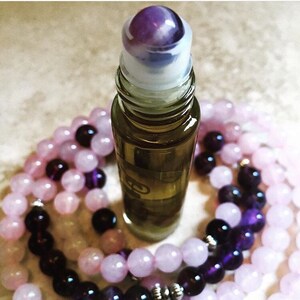 Peut inclure: Un petit flacon roll-on en verre vert olive avec une bille roulante en pierre précieuse violette est entouré de brins de perles de pierres précieuses roses et violettes. Les perles sont disposées en cercle autour du flacon, créant une esthétique de spa.