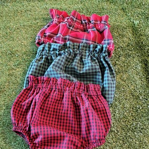 Può includere: Tre paia di bloomers per bambini in una pila. I bloomers sono realizzati in tessuto a quadri rosso, verde e rosso. Il tessuto ha un elastico in vita e aperture per le gambe. Lo sfondo è una superficie verde strutturata.