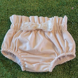 May include: A pale pink baby bloomer with a white lace bow. The bloomer is gathered at the waist and has a ruffled edge.