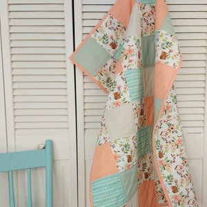 May include: A patchwork baby quilt with a floral and woodland animal print. The quilt is made of peach, mint green, and white fabric.