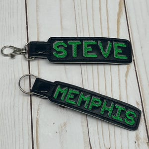 May include: Two black leather keychains with silver key rings. The keychains have green embroidered text. One keychain says "STEVE" and the other says "MEMPHIS".