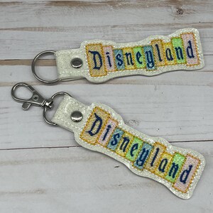 May include: Two white keychains with silver key rings. Each keychain has a colorful, glittery design with the word "Disneyland" in a vintage font.