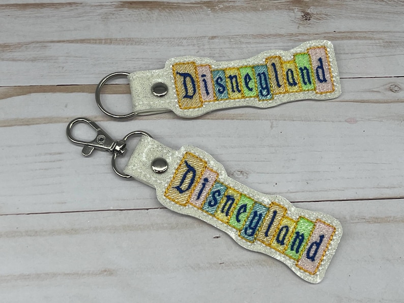 May include: Two white keychains with silver key rings. Each keychain has a colorful, glittery design with the word "Disneyland" in a vintage font.