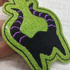 May include: Green glitter fabric keychain with a black silhouette of a character's horns with purple stitching.