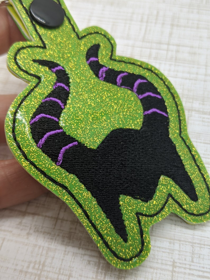 May include: Green glitter fabric keychain with a black silhouette of a character's horns with purple stitching.