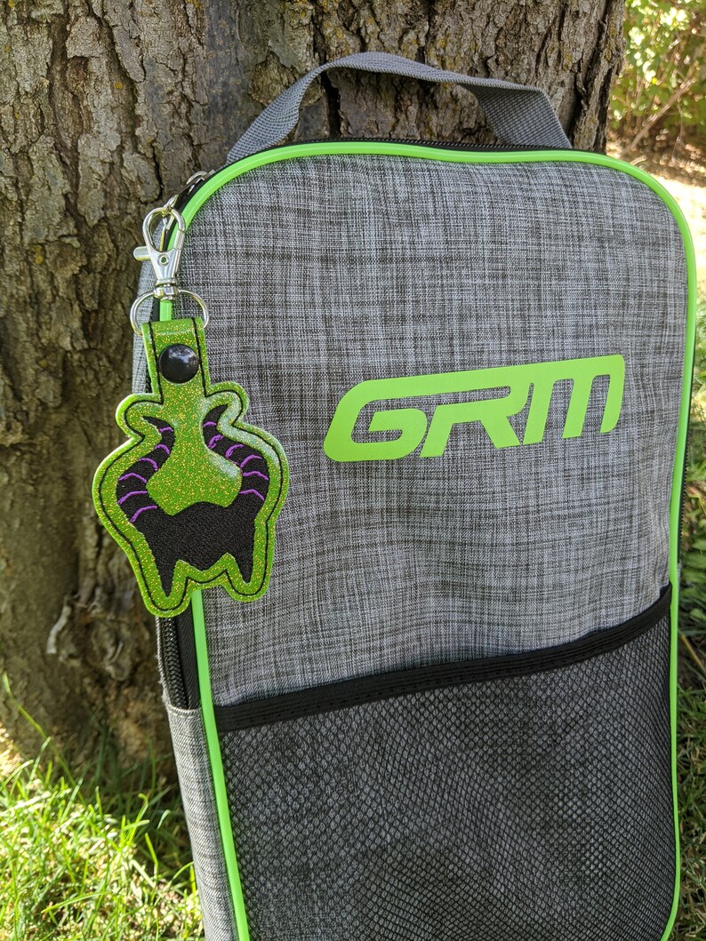 May include: A gray and green sports bag with a black mesh pocket and a green zipper. The bag has the word "GRIM" printed on the front in green letters. A green and purple monster keychain is attached to the bag.