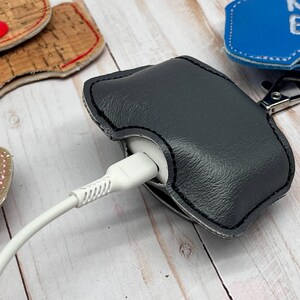 May include: A dark gray textured protective case for wireless earbuds, featuring black stitching and a metal lobster clasp. A white charging cable is plugged into the bottom of the white earbud case, which is partially enclosed by the gray cover.