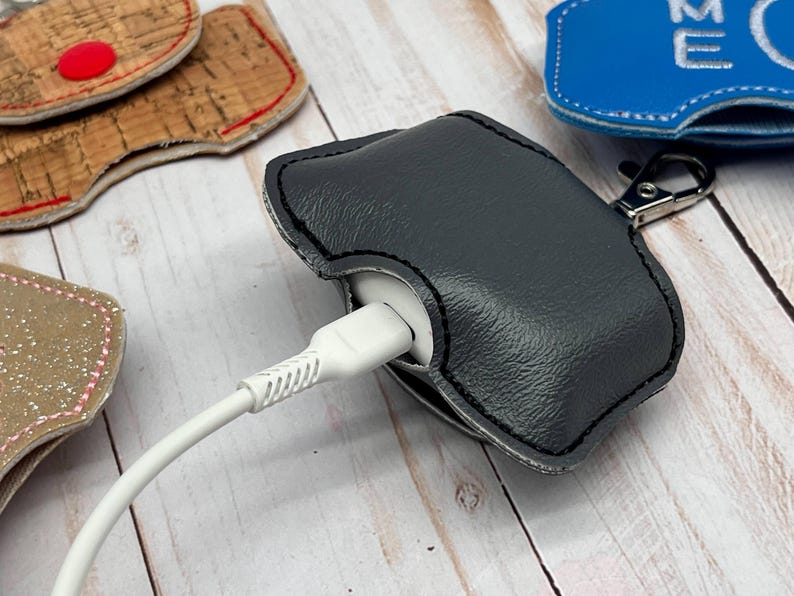 May include: A dark gray textured protective case for wireless earbuds, featuring black stitching and a metal lobster clasp. A white charging cable is plugged into the bottom of the white earbud case, which is partially enclosed by the gray cover.