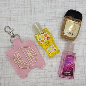 May include: A light pink faux leather hand sanitizer holder with a silver lobster clasp and embroidered monogram RJM, alongside a yellow Fresh Fruit hand sanitizer and a Bath & Body Works Warm Vanilla Sugar and a Berry Kissed hand sanitizer.
