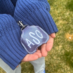 May include: A shield-shaped keychain with a shimmering purple surface and white embroidered letters reading ASC. The keychain features a silver-toned metal lobster clasp attached to the top, providing a secure way to fasten the item to accessories.