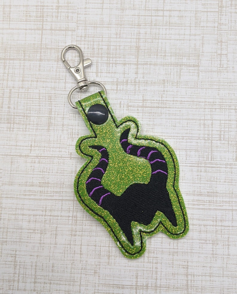 May include: Green glitter keychain with a black silhouette of the Evil Queen's horns from Snow White. The keychain has a silver metal clasp.