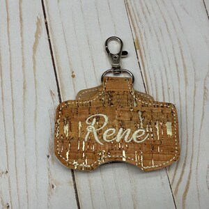 May include: A rectangular keychain holder made of natural cork material with metallic gold flecks throughout. The center features the name Rene embroidered in white cursive thread. A silver-toned metal lobster clasp is attached to the top loop.