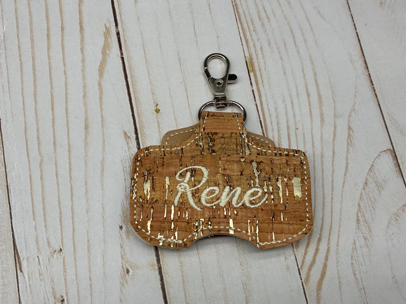 May include: A rectangular keychain holder made of natural cork material with metallic gold flecks throughout. The center features the name Rene embroidered in white cursive thread. A silver-toned metal lobster clasp is attached to the top loop.