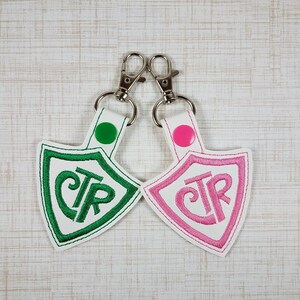 May include: Two shield-shaped keychains made of white material with silver metal lobster clasps. One keychain features green embroidery of the letters CTR and a green snap button. The other features pink embroidery of the letters CTR and a pink snap button.