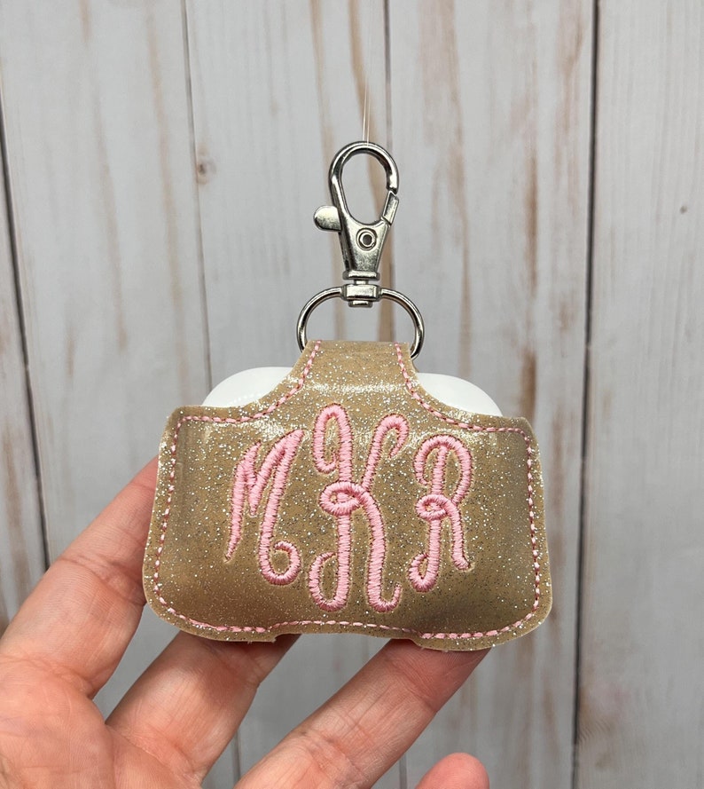 May include: A gold glittery protective case for wireless earbuds featuring a silver metal lobster clasp. The case is embroidered with the pink monogram letters MKR in a cursive script and has pink stitching along the edges of the material.