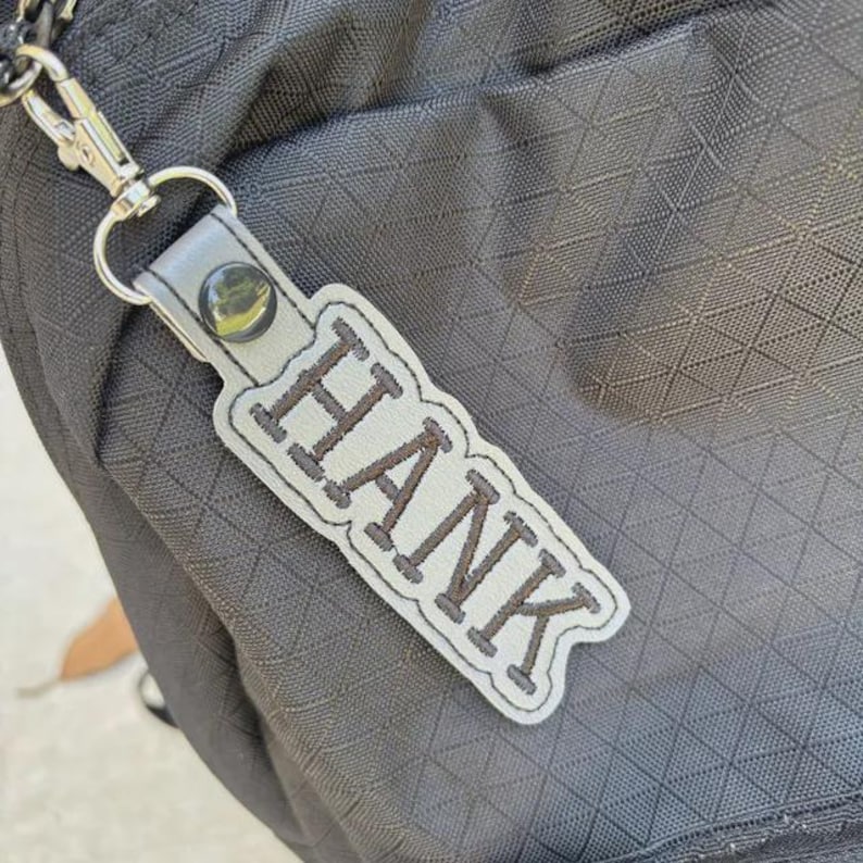Puede incluir: Un llavero de cuero gris con el nombre "HANK" bordado en letras marrones. El llavero est&aacute; sujeto a un cierre de metal plateado y se encuentra sobre un fondo negro texturizado.