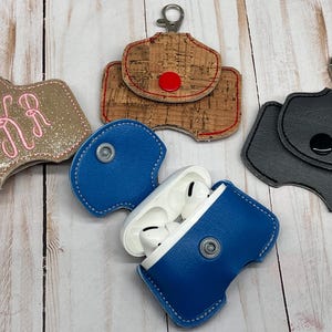 May include: Four protective cases for wireless earbuds, each featuring a metal lobster clasp. One case is gold glitter with pink monogram embroidery, one is cork with a red snap, one is solid black with a black snap, and one is blue with a silver snap.