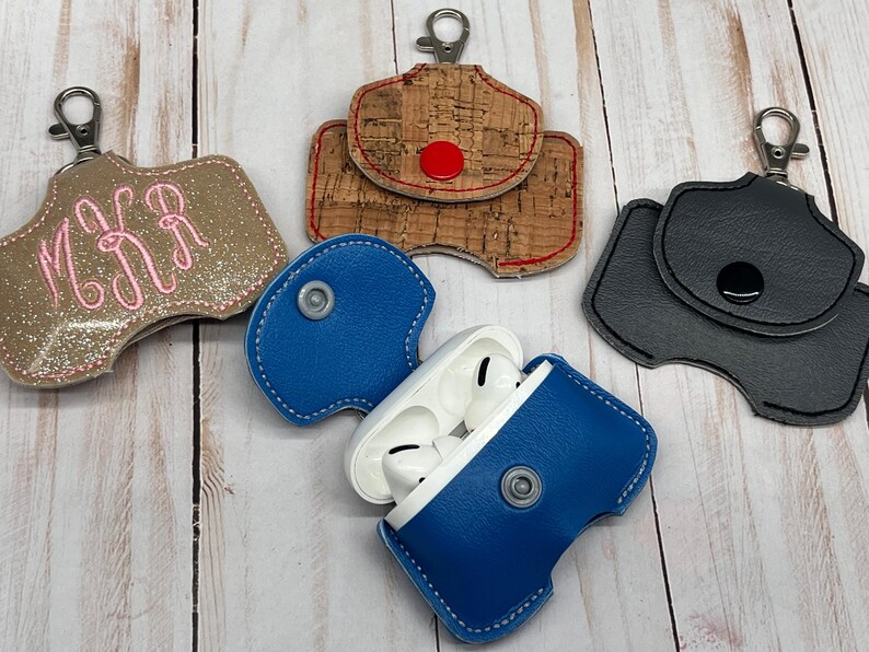 May include: Four protective cases for wireless earbuds, each featuring a metal lobster clasp. One case is gold glitter with pink monogram embroidery, one is cork with a red snap, one is solid black with a black snap, and one is blue with a silver snap.