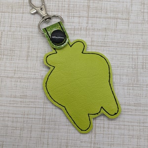 May include: A green felt keychain with a black stitched outline in the shape of a cartoon monster. The keychain has a silver metal clasp and a green glitter strap with a black button.