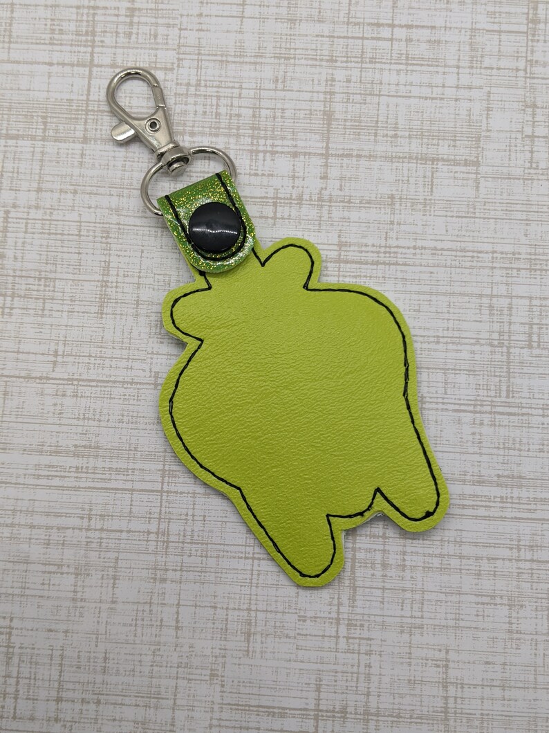 May include: A green felt keychain with a black stitched outline in the shape of a cartoon monster. The keychain has a silver metal clasp and a green glitter strap with a black button.