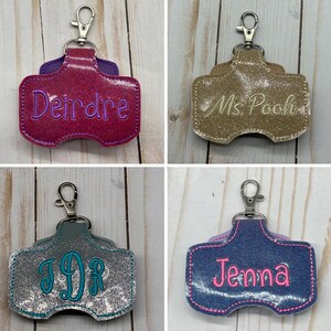 May include: Four glittery, rectangular keychain holders with lobster claw clasps. The top left is magenta with purple text reading Deirdre. The top right is gold with white text reading Ms. Pooh. The bottom left is silver with teal monogram text JDR. The bottom right is blue with pink text reading Jenna.