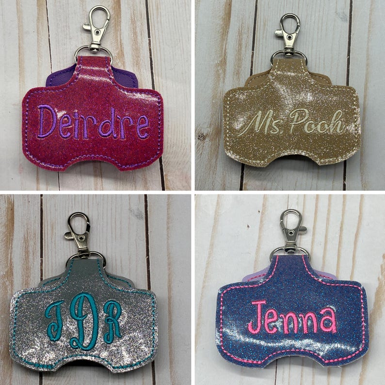 May include: Four glittery, rectangular keychain holders with lobster claw clasps. The top left is magenta with purple text reading Deirdre. The top right is gold with white text reading Ms. Pooh. The bottom left is silver with teal monogram text JDR. The bottom right is blue with pink text reading Jenna.
