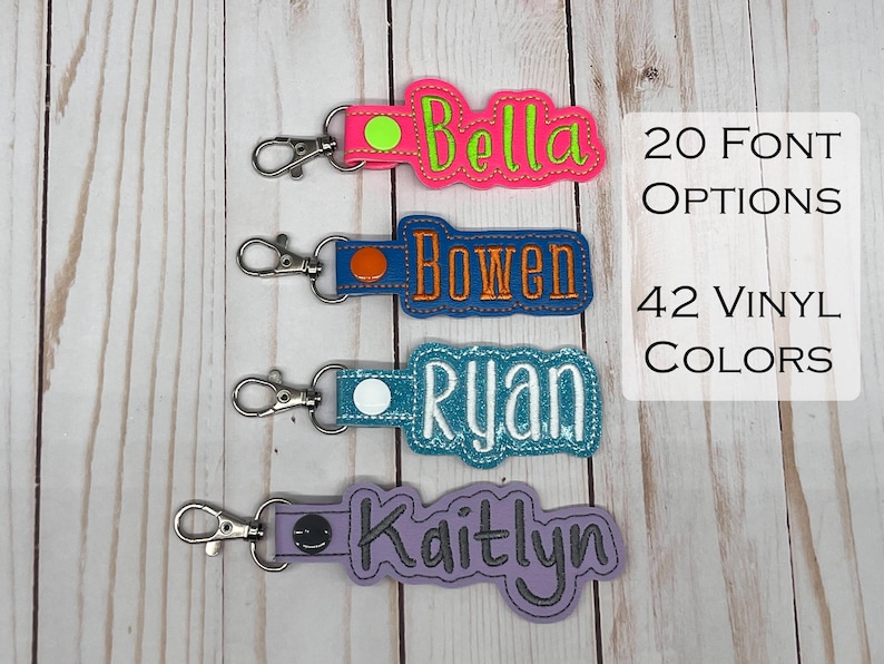 Puede incluir: Cuatro llaveros personalizados con nombres: "Bella" en rosa y verde, "Bowen" en azul y naranja, "Ryan" en turquesa y blanco, y "Kaitlyn" en morado y gris. Cada llavero tiene un cierre plateado. El texto a la derecha dice "20 Opciones de fuente" y "42 Colores de vinilo."