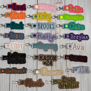 Puede incluir: Una colecci&oacute;n de llaveros bordados en varios colores, cada uno con un nombre o palabra. Los llaveros tienen un cierre de metal y est&aacute;n dispuestos sobre una superficie de madera clara. Los nombres incluyen Lexi, Whitney, Bella, y otros.