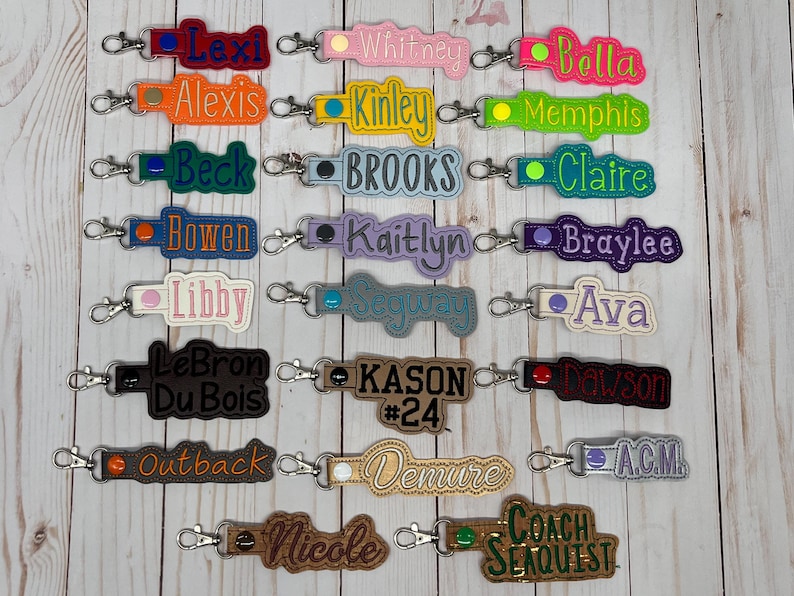 Puede incluir: Una colecci&oacute;n de llaveros bordados en varios colores, cada uno con un nombre o palabra. Los llaveros tienen un cierre de metal y est&aacute;n dispuestos sobre una superficie de madera clara. Los nombres incluyen Lexi, Whitney, Bella, y otros.