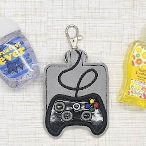 May include: A gray and black video game controller keychain with a loop for attaching to a bag or belt. The keychain has a pocket for a small bottle of hand sanitizer. The hand sanitizer bottle is clear with a yellow label and a design of citrus fruits. The text on the label reads "Hand Sanitizer Fresh Fruit Scented 1 fl oz US (29.6 ml)".
