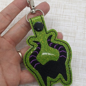May include: Green glitter keychain with a black silhouette of Maleficent's horns with purple accents.