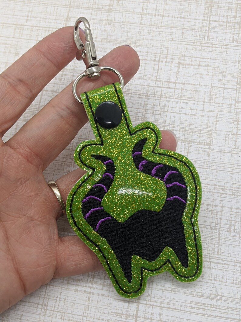 May include: Green glitter keychain with a black silhouette of Maleficent's horns with purple accents.