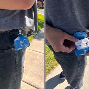 May include: A bright blue leather case for wireless earbuds featuring a silver metal snap closure and a silver keychain clip. The front of the case is embossed with the letters M and C stacked vertically in a white font.