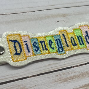 May include: A white keychain with a colorful embroidered design that says "Disneyland".