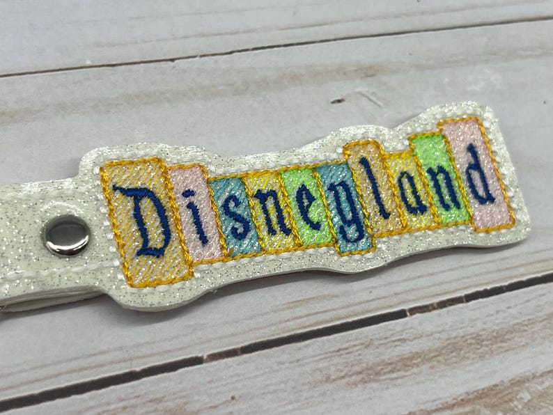 May include: A white keychain with a colorful embroidered design that says "Disneyland".