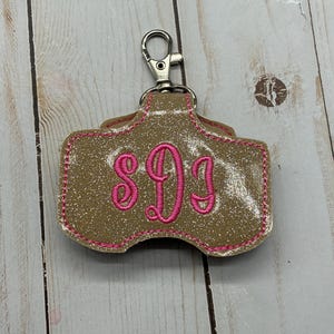 May include: A glittery gold keychain holder featuring a silver metal lobster clasp at the top. The holder has a rounded rectangular shape with pink embroidered stitching along the edges and a pink embroidered monogram of the letters SDJ.