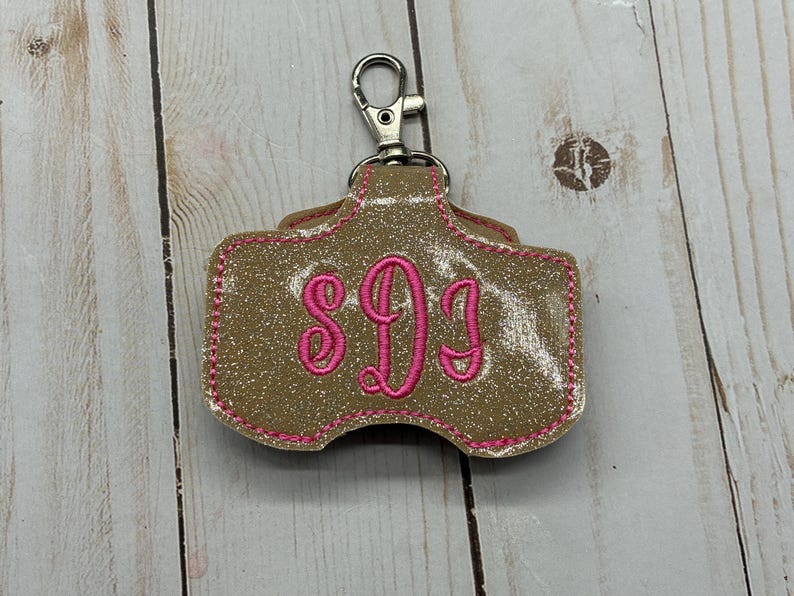 May include: A glittery gold keychain holder featuring a silver metal lobster clasp at the top. The holder has a rounded rectangular shape with pink embroidered stitching along the edges and a pink embroidered monogram of the letters SDJ.